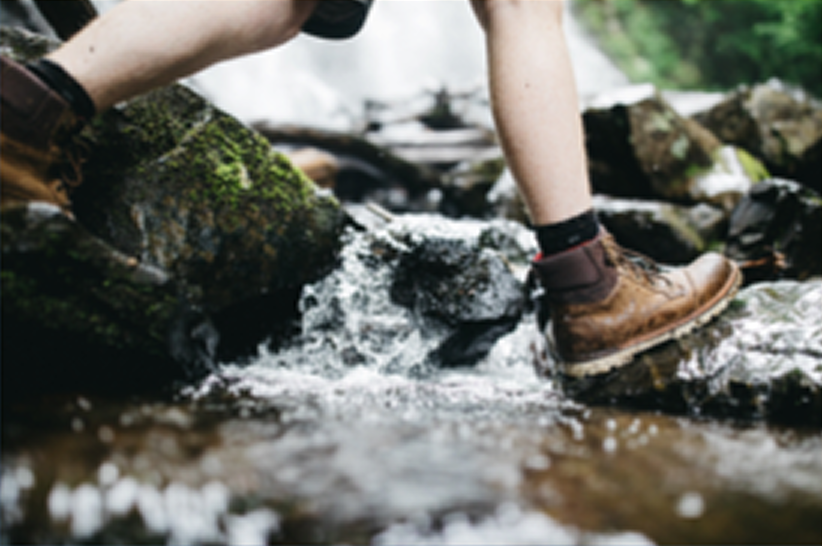 Person wearing brown boots steps on rocks across a shallow stream with running water; green moss is visible on some rocks in the background.