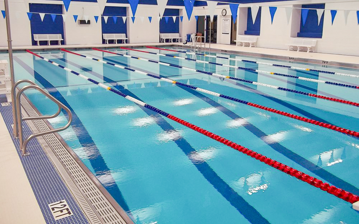 Indoor swimming pool with lane dividers, blue and white wall banners, pool ladders, white benches, and a depth marker showing 12 feet. The pool area is empty.