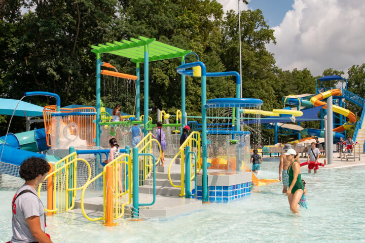 People enjoy a colorful outdoor water park with slides, splash features, and shallow water on a sunny day; trees are visible in the background.