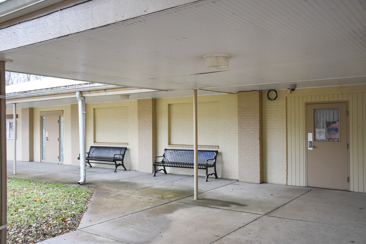 Covered walkway with two black metal benches against a beige brick wall and two closed doors in the background.