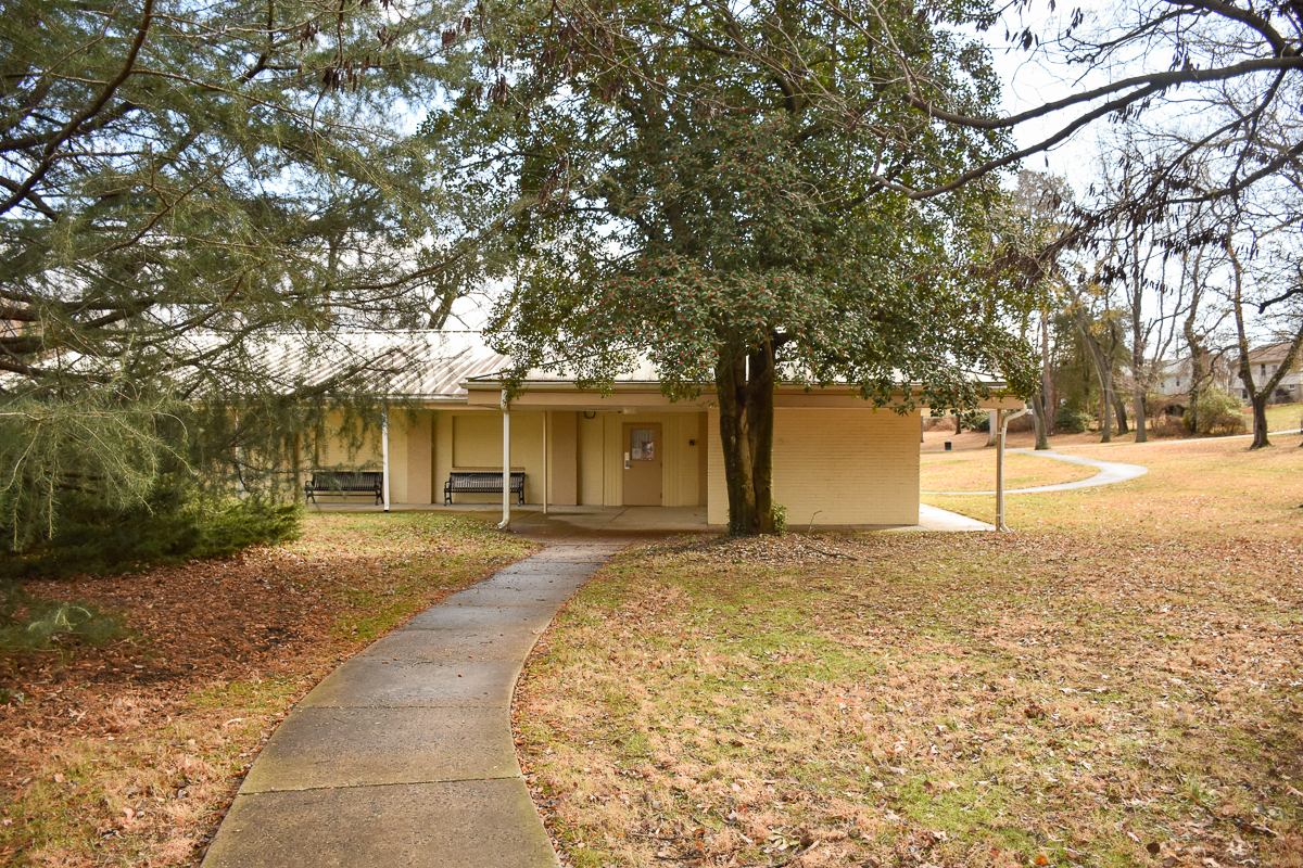 A concrete path leads to a small beige building with a covered porch, surrounded by grass, trees, and leaf litter on the ground.