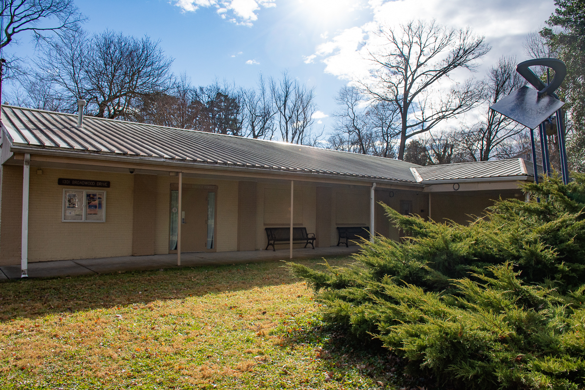 Single-story beige brick building with a metal roof, notice boards, benches, and nearby bushes, under a sunny sky with bare trees in the background.