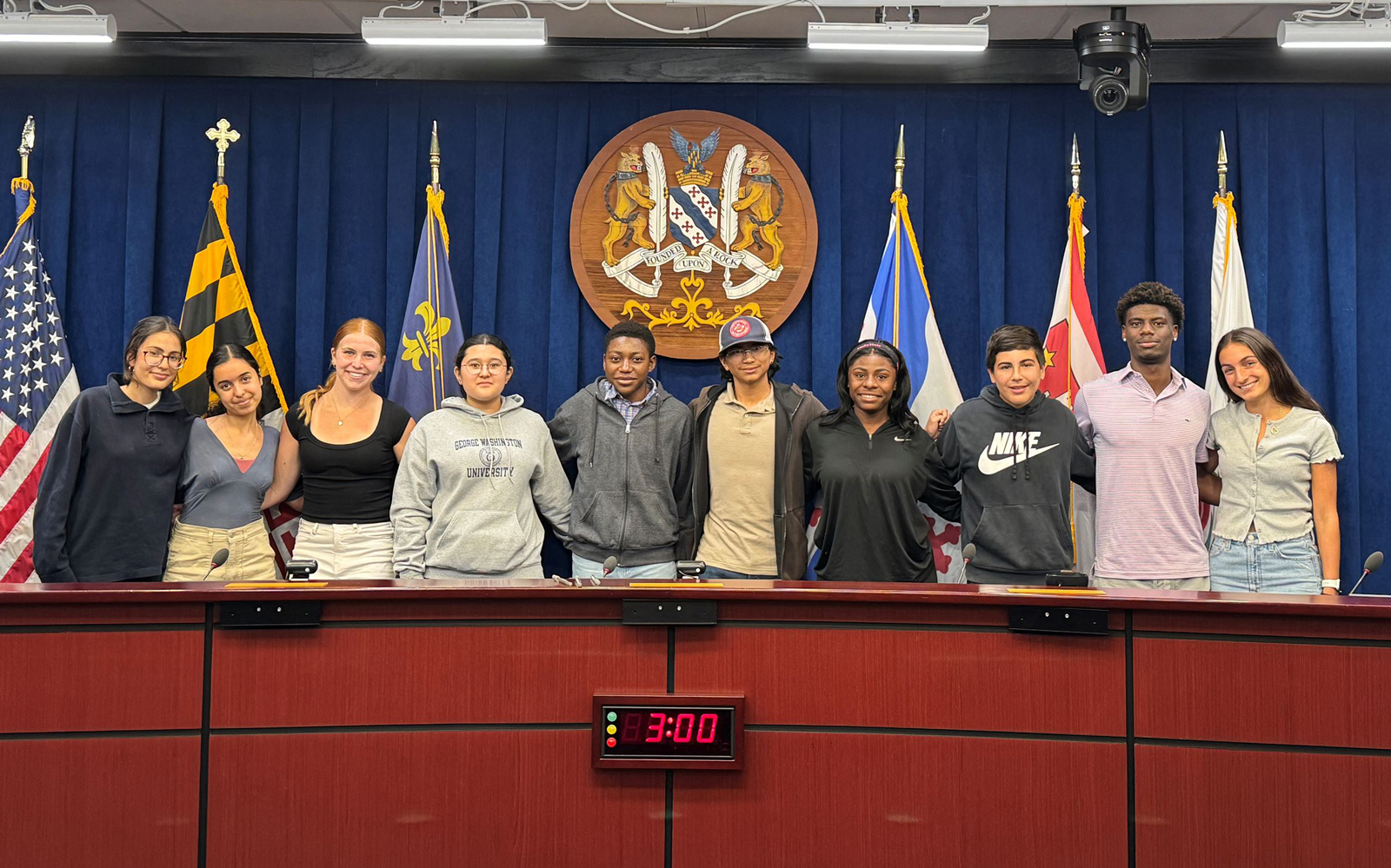 Youth-Commission-2025 Ten young people stand side by side behind a council desk with flags and a seal in the background; a digital clock displays