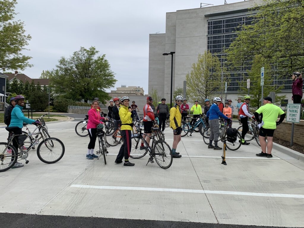 A group of cyclists with bikes gather in a parking lot on a cloudy day, standing and talking near a modern office building.