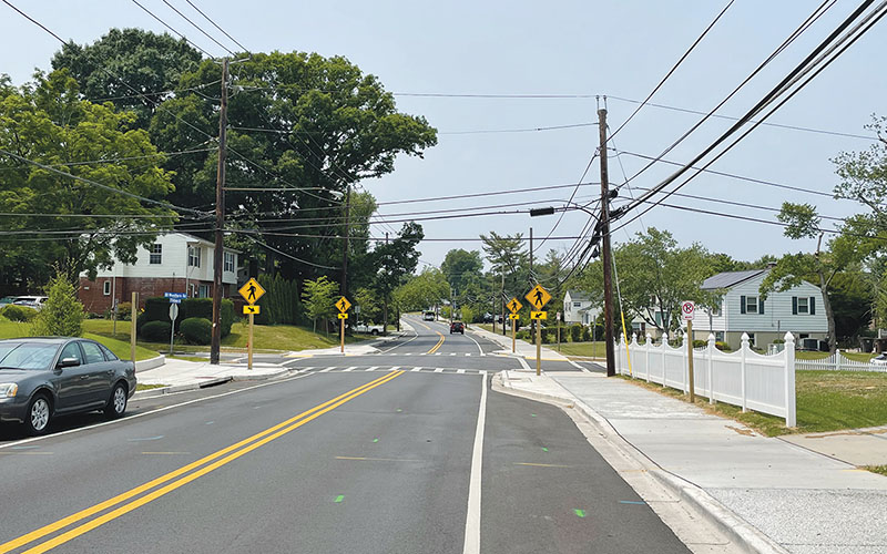 A two-lane suburban road with yellow lane markings, crosswalk, warning signs, parked car, houses, and overhead utility lines on a clear day.