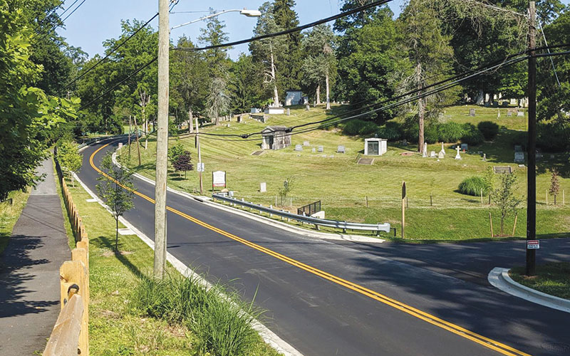A paved road with double yellow lines curves alongside a cemetery on a grassy hill, bordered by trees, utility poles, and a sidewalk with a wooden fence.