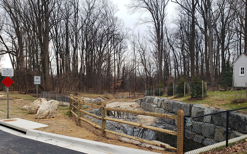A fenced drainage area with rocks runs next to a small house and leafless trees on an overcast day; signs and a sidewalk are visible in the foreground.