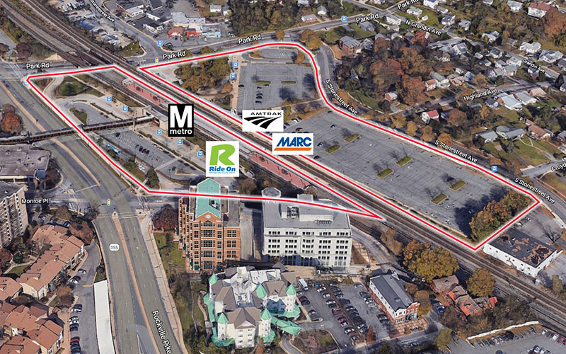 Aerial view of a large parking lot bordered in red, with logos for Metro, Amtrak, MARC, and Ride On transit services, surrounded by roads, buildings, and train tracks.