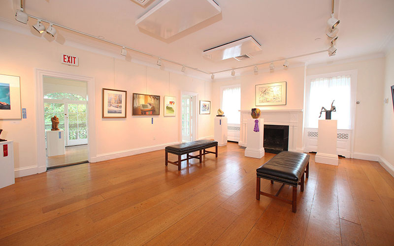 A well-lit art gallery room with wooden floors, benches, paintings on the walls, and sculptures on pedestals, with large windows and an exit door visible.