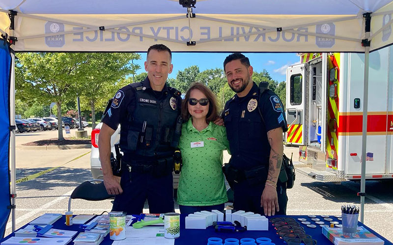 Two police officers and a woman stand under a "Rockville City Police" tent at an outdoor event, with information materials displayed on a table in front of them.