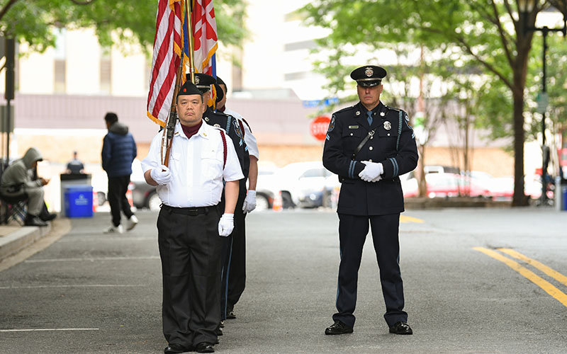 A uniformed color guard holds flags while another officer stands nearby on a city street during a formal outdoor event.