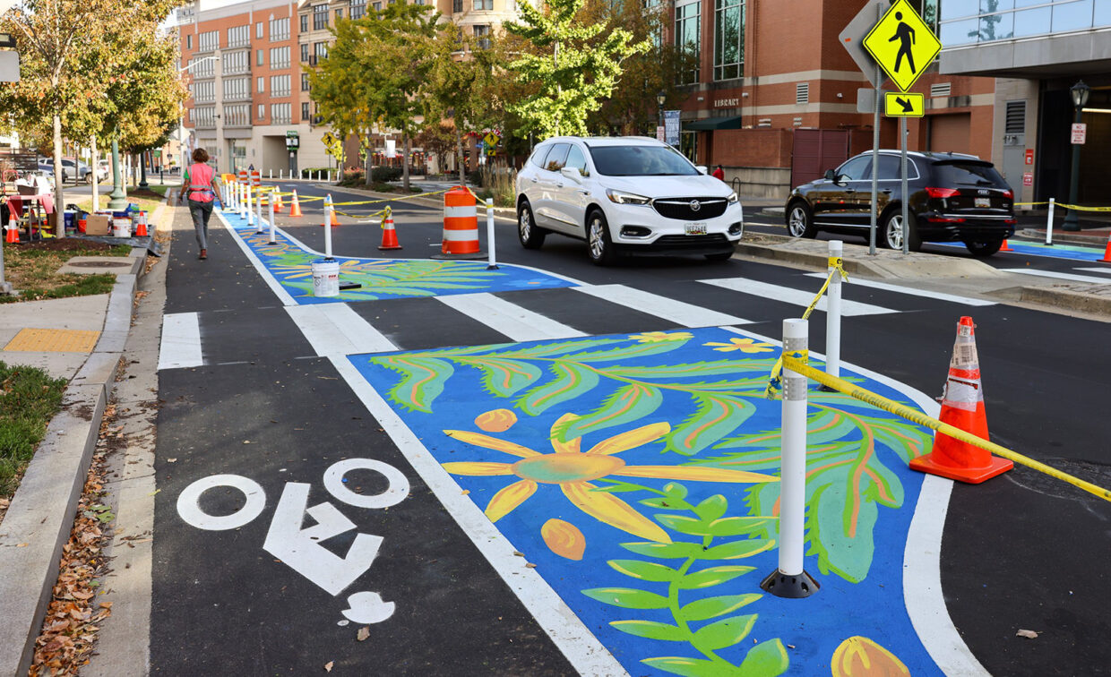 A city street with bright floral murals painted on the road, bike lane markings, traffic cones, cars, and pedestrians nearby.