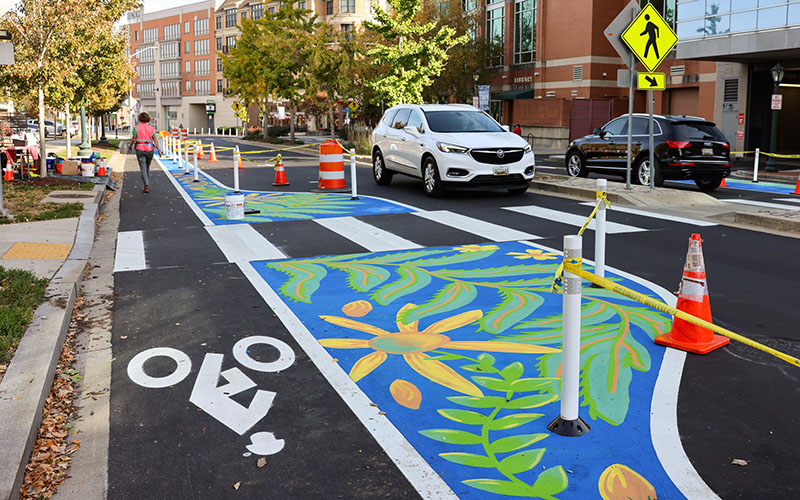A street crosswalk is painted with colorful floral designs. Traffic cones and caution tape mark the area, and two cars wait near the crossing.