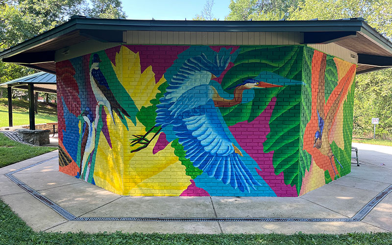 A colorful mural of large birds, including herons and parrots, painted on the exterior brick wall of a pavilion in a park setting.