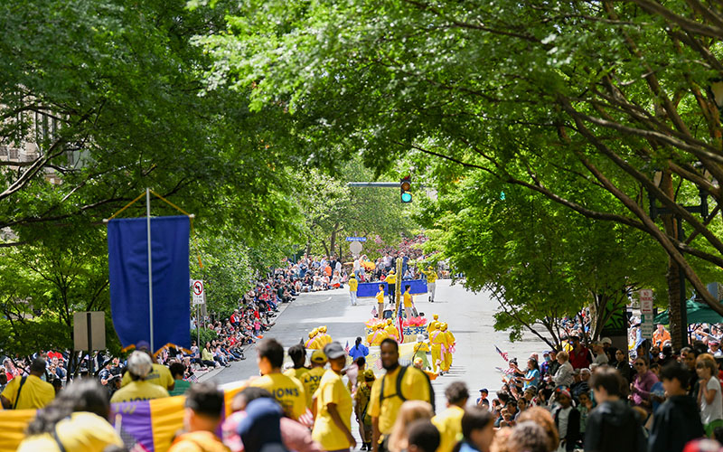 A parade moves down a tree-lined street with participants in yellow costumes, watched by crowds seated along both sides of the road.