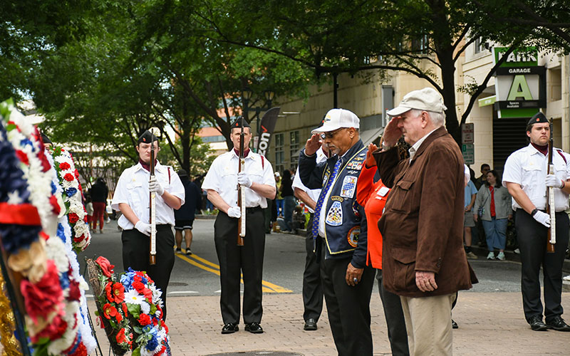 People stand and salute in front of wreaths at a memorial event, while uniformed individuals with rifles stand in formation on a city street.