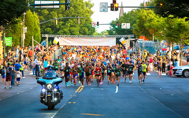 A large group of runners begins a race on a city street, with a police officer on a motorcycle leading the way and spectators watching from the sidelines.