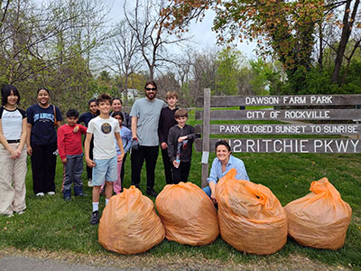 A group of people pose by large orange trash bags in front of a Dawson Farm Park sign, after collecting litter. Trees and grass are visible in the background.