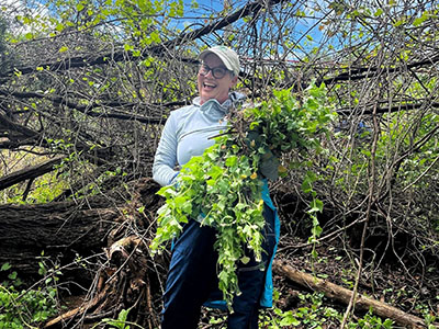 A person wearing glasses and a cap stands outdoors, smiling and holding a large bundle of green plants in front of fallen branches and trees.