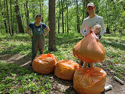 Two adults and a child stand in a wooded area with four large orange trash bags filled with collected litter.