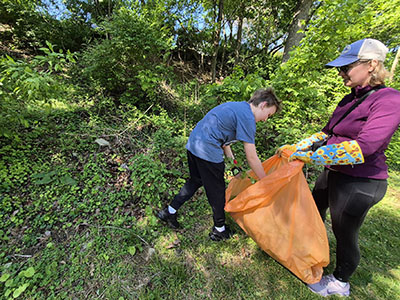 A woman and a child pick up litter and place it in an orange bag while standing on grass near dense green foliage.
