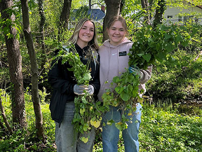 Two people stand outdoors in a wooded area, smiling and holding large bundles of pulled-up plants or weeds.