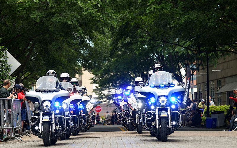 A group of police officers on motorcycles with flashing lights rides down a city street lined with spectators and trees.