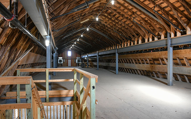 Spacious attic with exposed wooden beams, unfinished walls, and ceiling lights; a wooden railing and staircase are visible in the foreground.