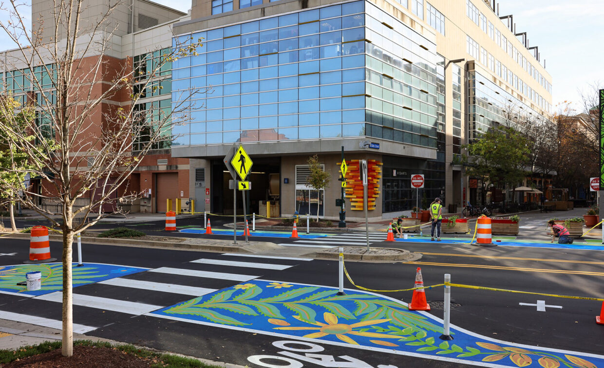 A city street with colorful painted crosswalks and traffic barriers, near a modern glass building; workers and traffic cones are present.