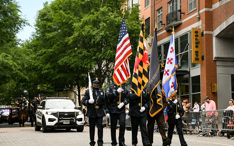 A color guard in uniform marches in a parade, holding American, Maryland, and other flags, with a police car and spectators in the background.