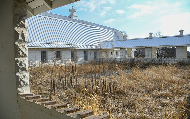 Courtyard with overgrown dry grass surrounded by a white stone building with a metal roof under a partly cloudy sky.