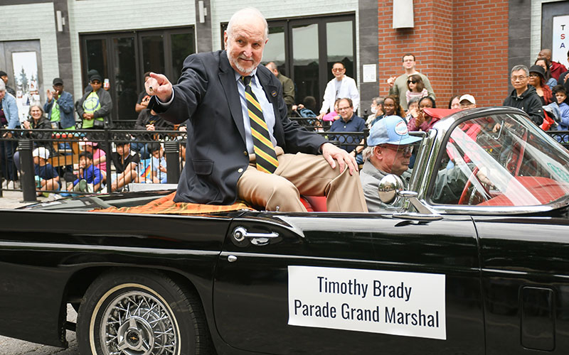 An older man in a suit sits atop the back of a convertible car labeled 