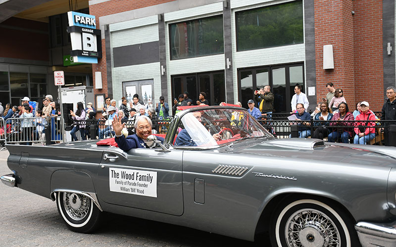 A person waves from a gray Ford Thunderbird convertible in a parade, with a sign reading 