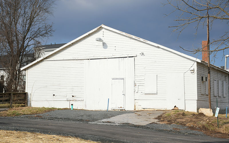 A white, weathered barn with a large sliding door and a small entry door stands on a gravel and dirt lot under a cloudy sky.