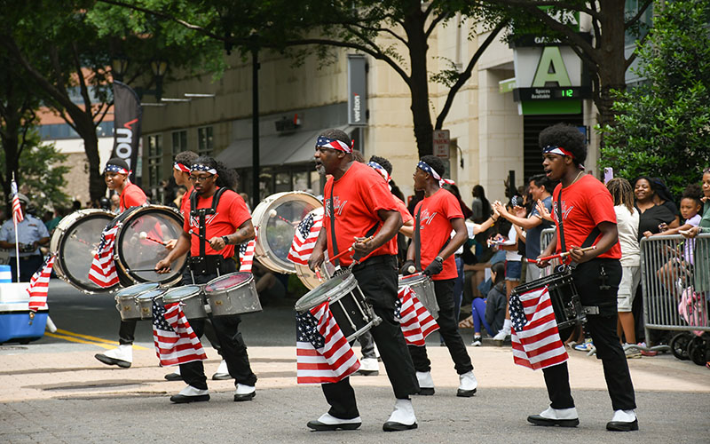 A group of drummers in red shirts and headbands perform in a parade, with American flags on their drums and spectators watching from the sidewalk.