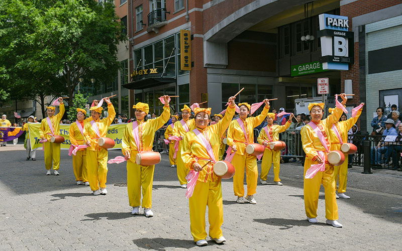 A group of performers in yellow costumes play waist drums and smile while marching in a parade on a city street.