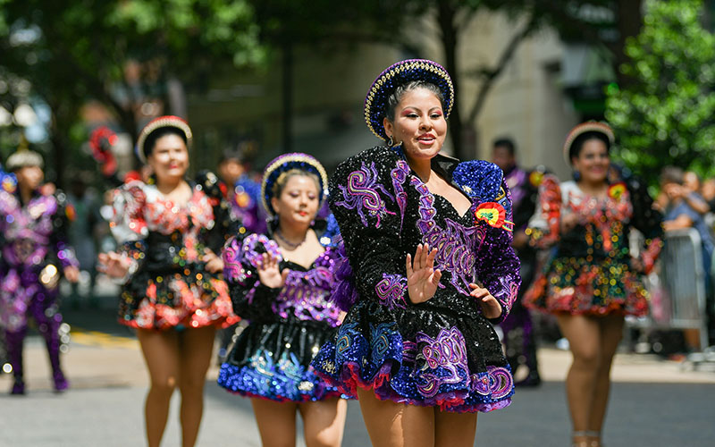 A group of women in colorful, sequined costumes dance in a street parade during daylight, with trees and spectators in the background.