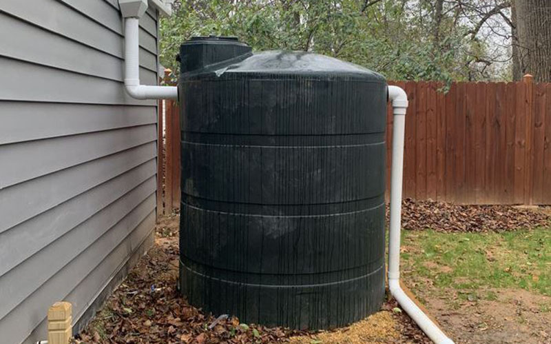 A large black rainwater collection tank is connected to a house with white PVC pipes, positioned near a wooden fence and surrounded by fallen leaves.