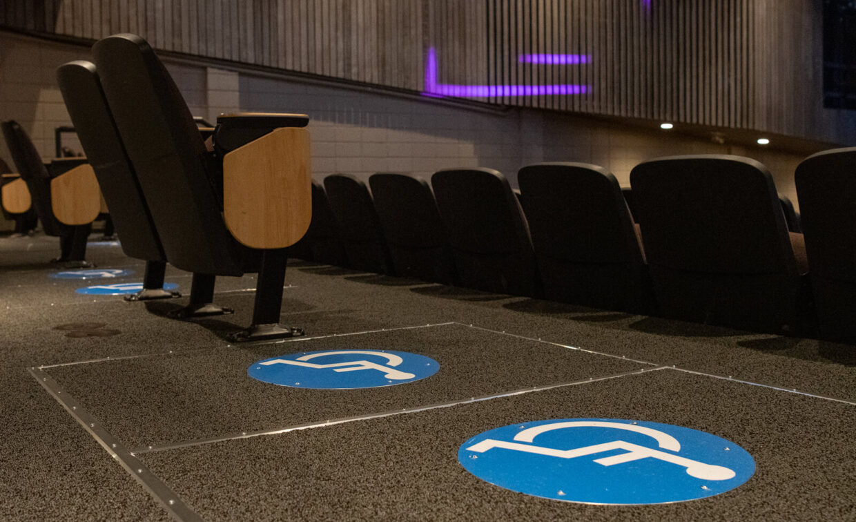 Several theater seats with wheelchair-accessible spaces marked by blue signs are shown inside an auditorium.