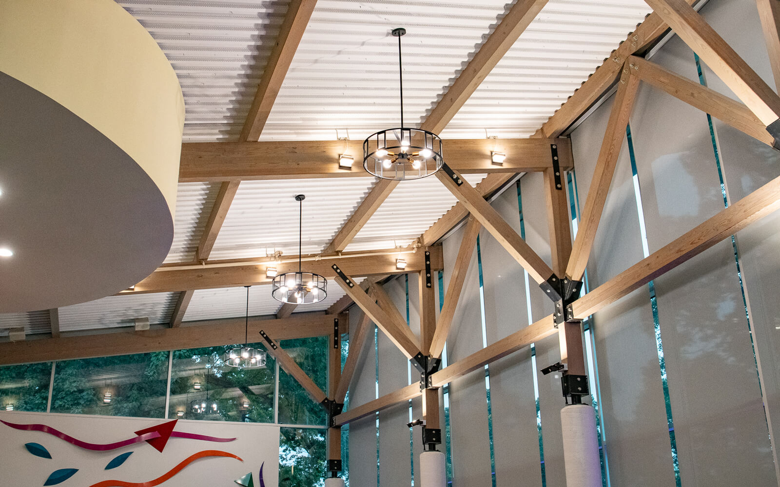 Interior ceiling with exposed wooden beams, metal supports, and modern hanging light fixtures; large windows on the right let in natural light.