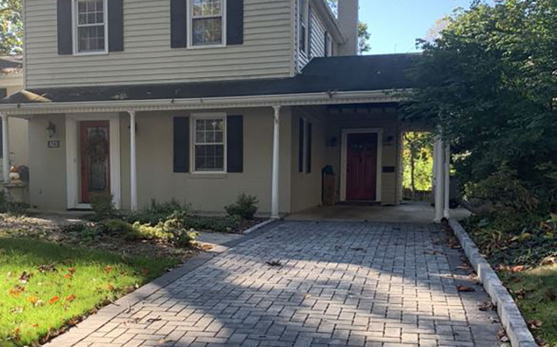 A two-story house with light-colored siding, black shutters, a covered front porch, and a paved driveway leading to a carport.