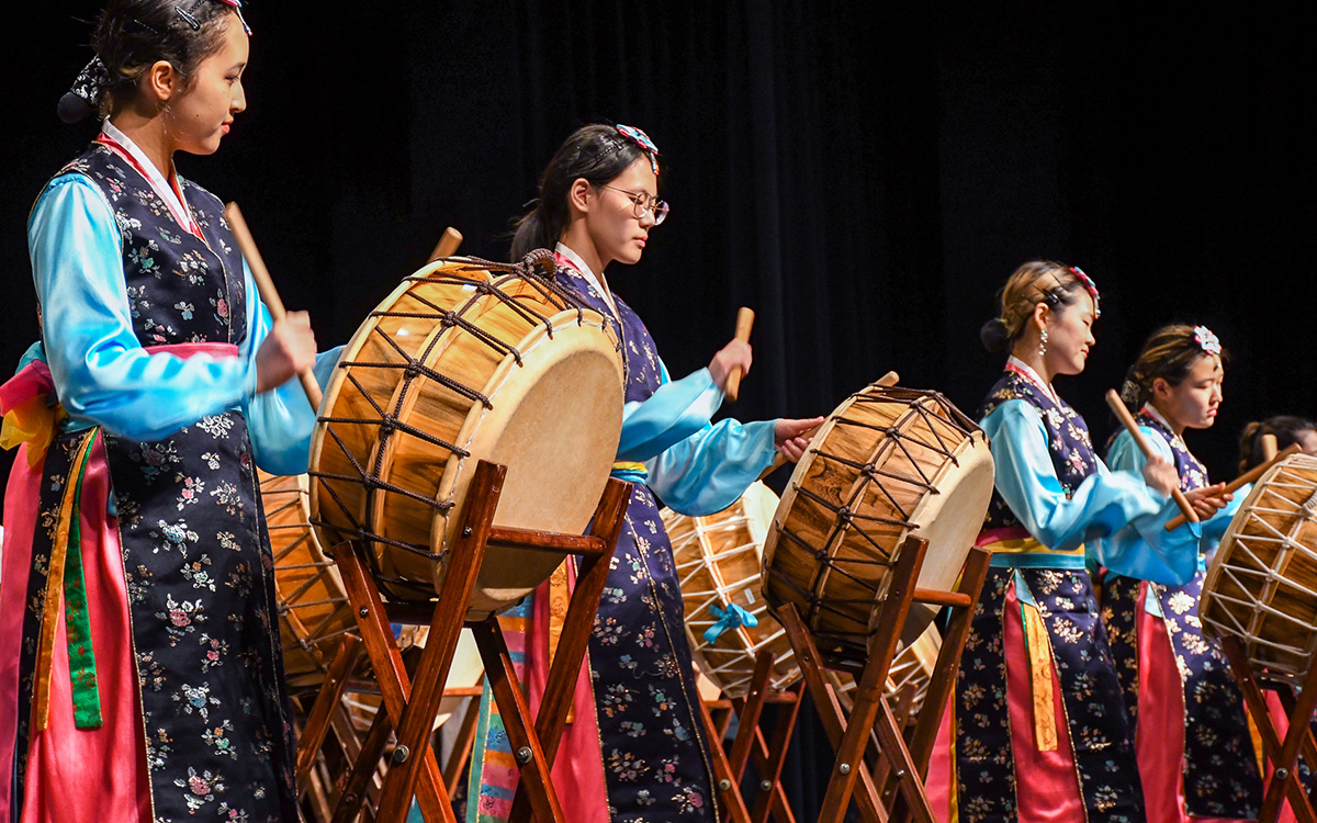 Five women in traditional Korean clothing play large barrel drums on stage, each holding a pair of drumsticks, performing in a coordinated line.