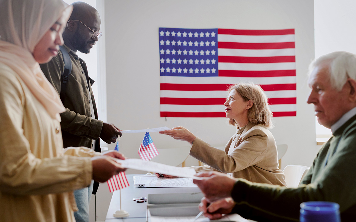 People at a table participate in a civic event with paperwork; a U.S. flag is displayed on the wall in the background. People at a table participate in a civic event with paperwork; a U.S. flag is displayed on the wall in the background.