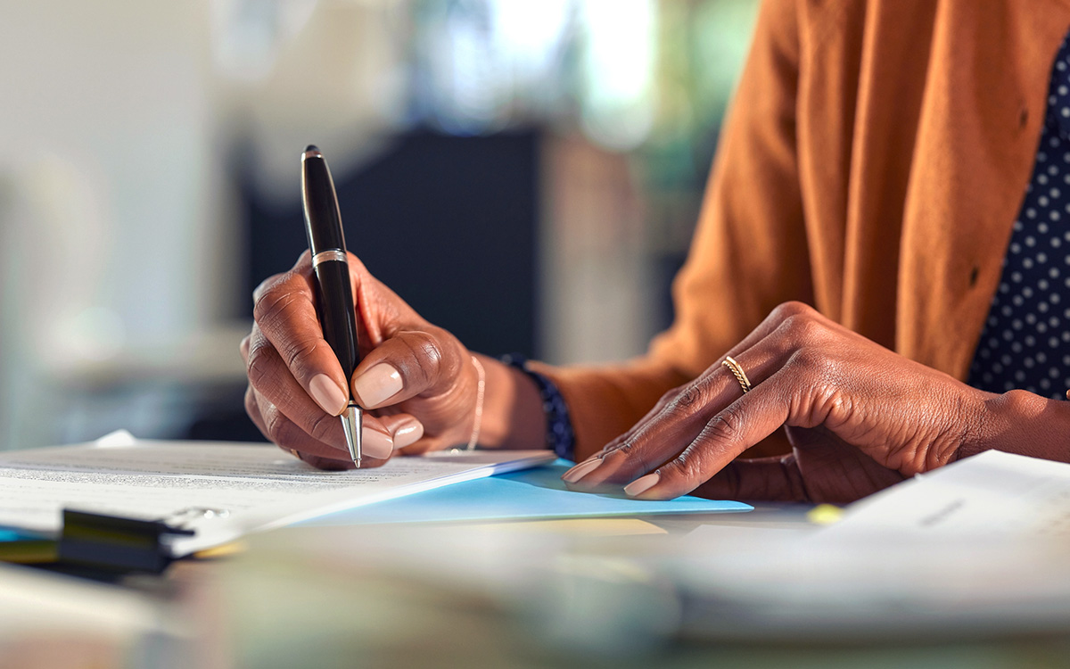 A person wearing a brown jacket is writing on a document with a pen at a desk, surrounded by papers and folders.