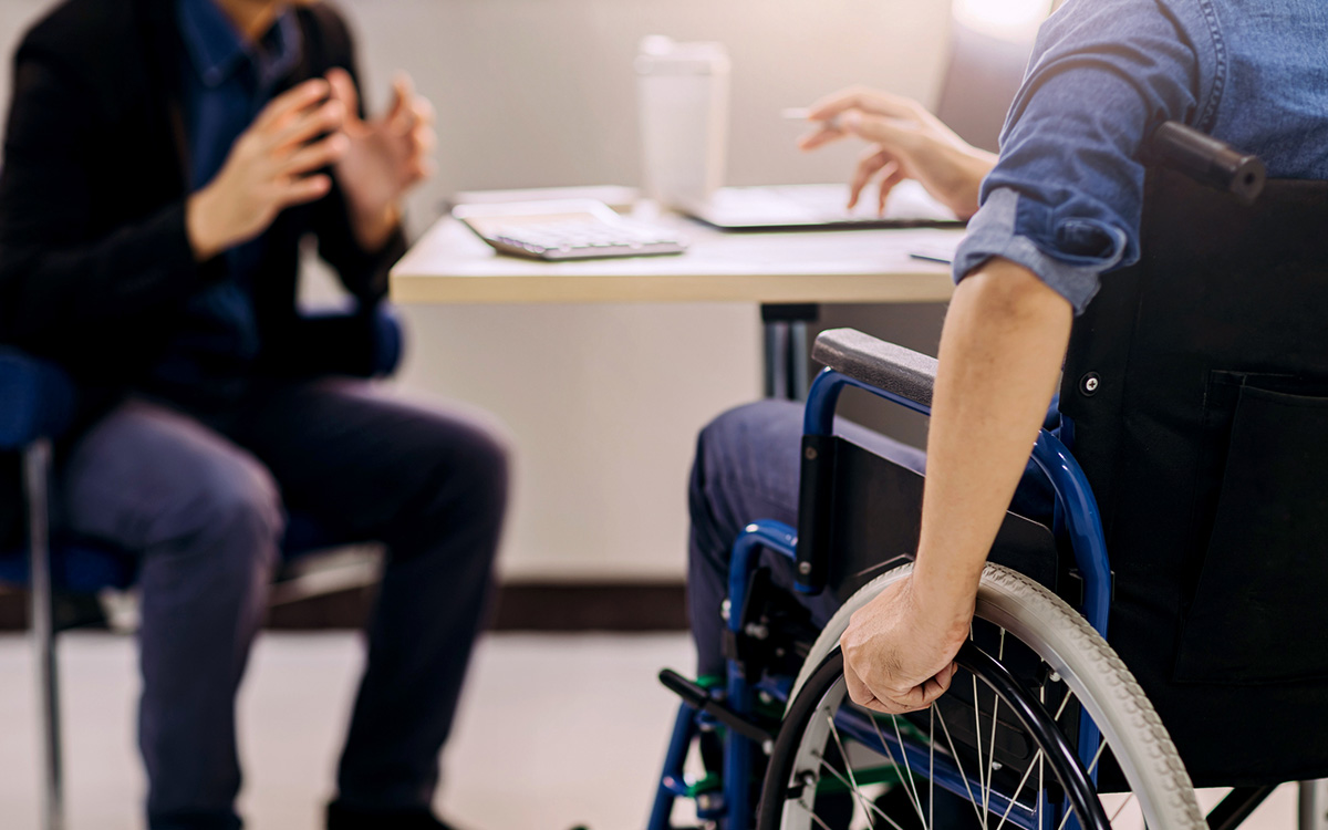 Two people are having a discussion at a desk; one person is seated in a wheelchair, and only their hands and part of their upper bodies are visible.