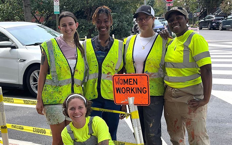 Five women in construction attire stand and kneel beside a caution sign that reads "WOMEN WORKING" on a city sidewalk, with traffic and trees in the background.