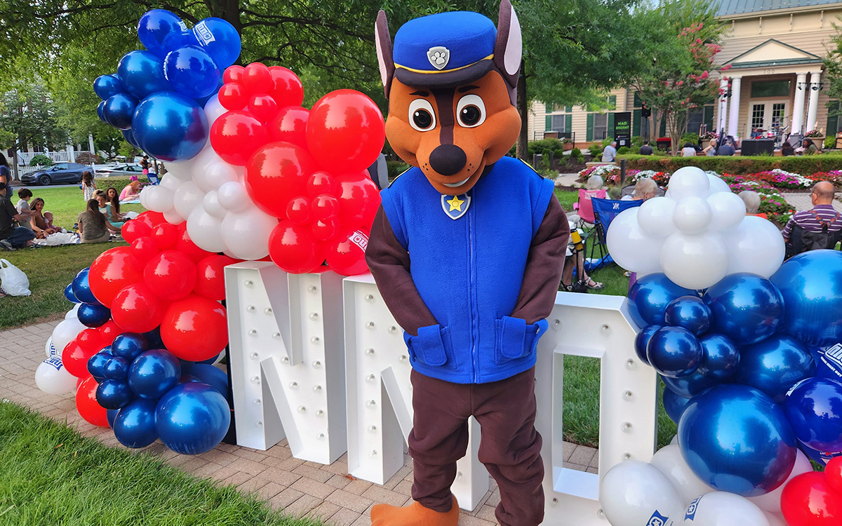A person in a canine mascot costume stands in front of red, white, and blue balloons and large  A person in a canine mascot costume stands in front of red, white, and blue balloons and large