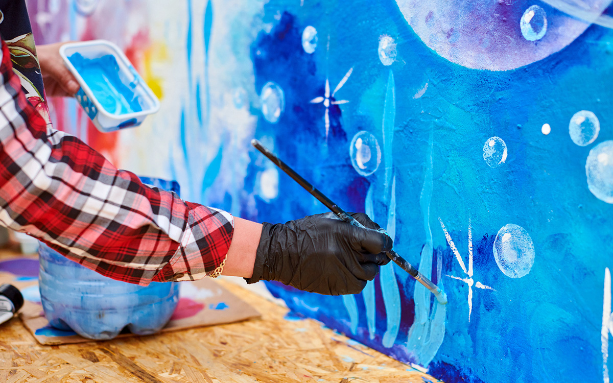 A person wearing a plaid shirt and black gloves paints blue and white shapes on a colorful mural, holding a paintbrush and tray. A person wearing a plaid shirt and black gloves paints blue and white shapes on a colorful mural, holding a paintbrush and tray.