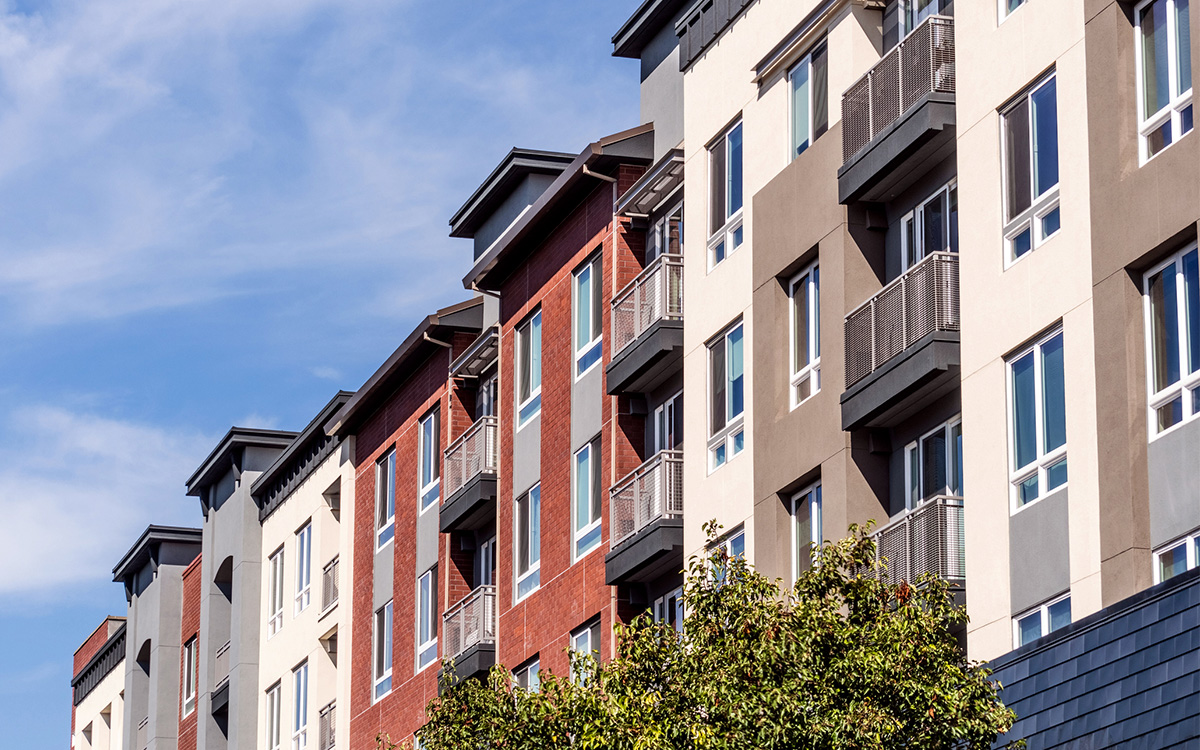 Modern multi-story apartment building with balconies, various exterior colors, and a tree in the foreground under a blue sky. Modern multi-story apartment building with balconies, various exterior colors, and a tree in the foreground under a blue sky.