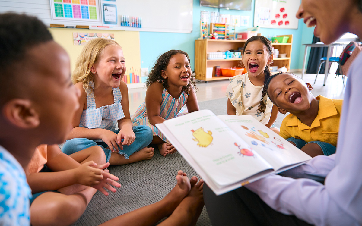 A group of young children sit in a circle on the classroom floor, smiling and laughing as an adult reads a picture book to them.